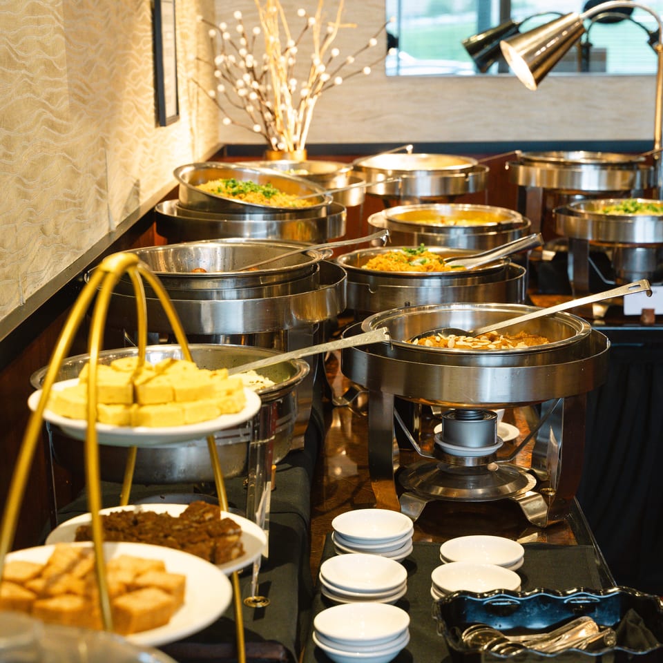 Elegant restaurant buffet with stainless-steel chafing dishes, plated desserts, serving utensils, and warm decorative lighting.