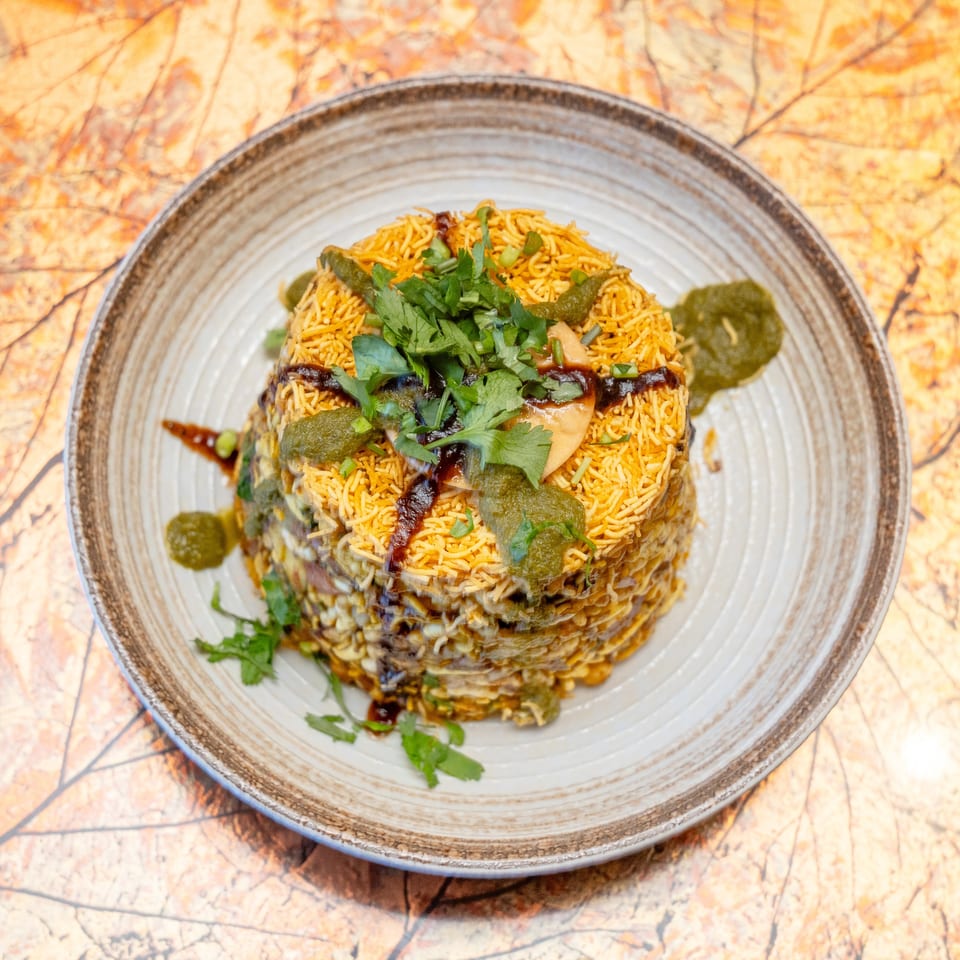 Layered Indian rice dish topped with sev, cilantro, green chutney, and dark tamarind sauce on a decorative plate.