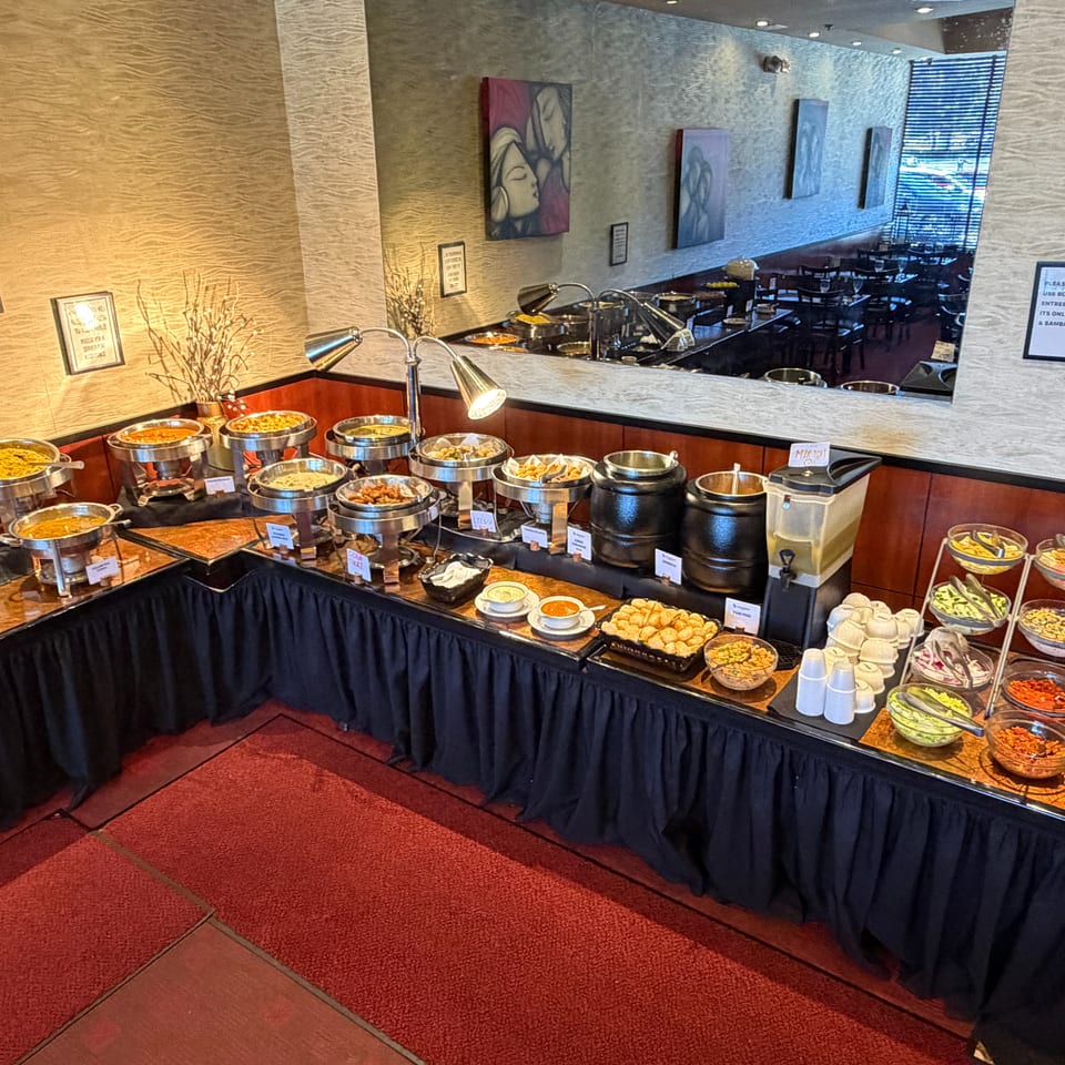 Indian restaurant buffet featuring chafing dishes, colorful curries, sauces, salads, desserts, and condiments in a warmly decorated dining room.