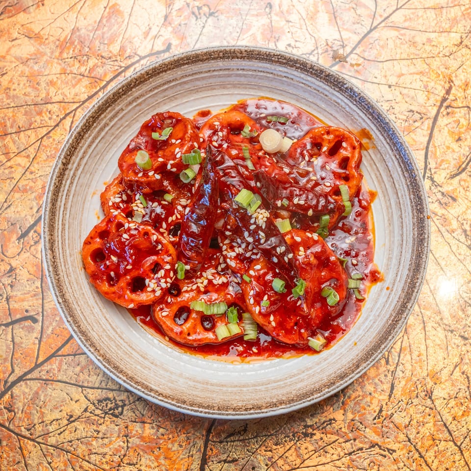 Plate of lotus root slices in glossy red chili sauce, garnished with sesame seeds and green onions on a patterned tabletop.