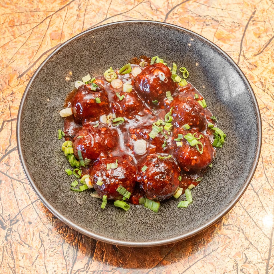 Glazed meatballs topped with chopped green onions in a dark ceramic bowl on a marbled tabletop.