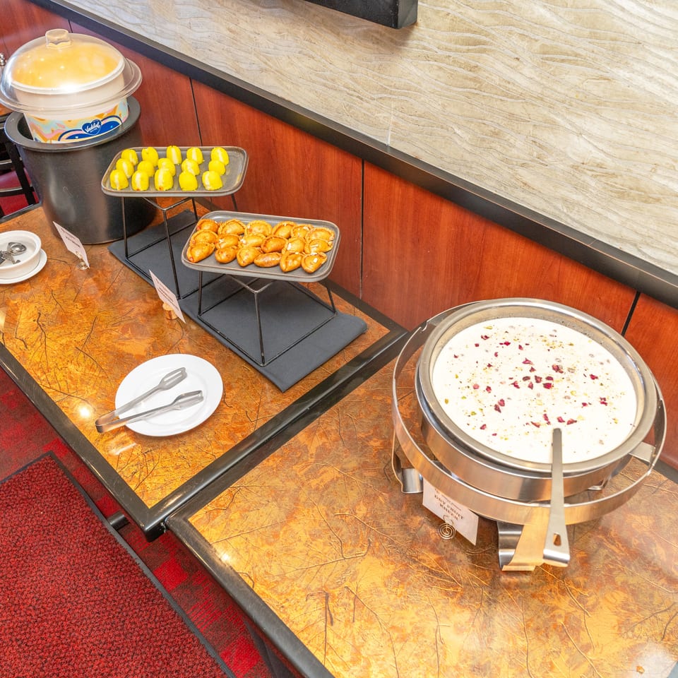 Restaurant buffet displays yellow pastries, golden fried snacks, creamy dessert, serving utensils, and a covered dish on tiered trays.