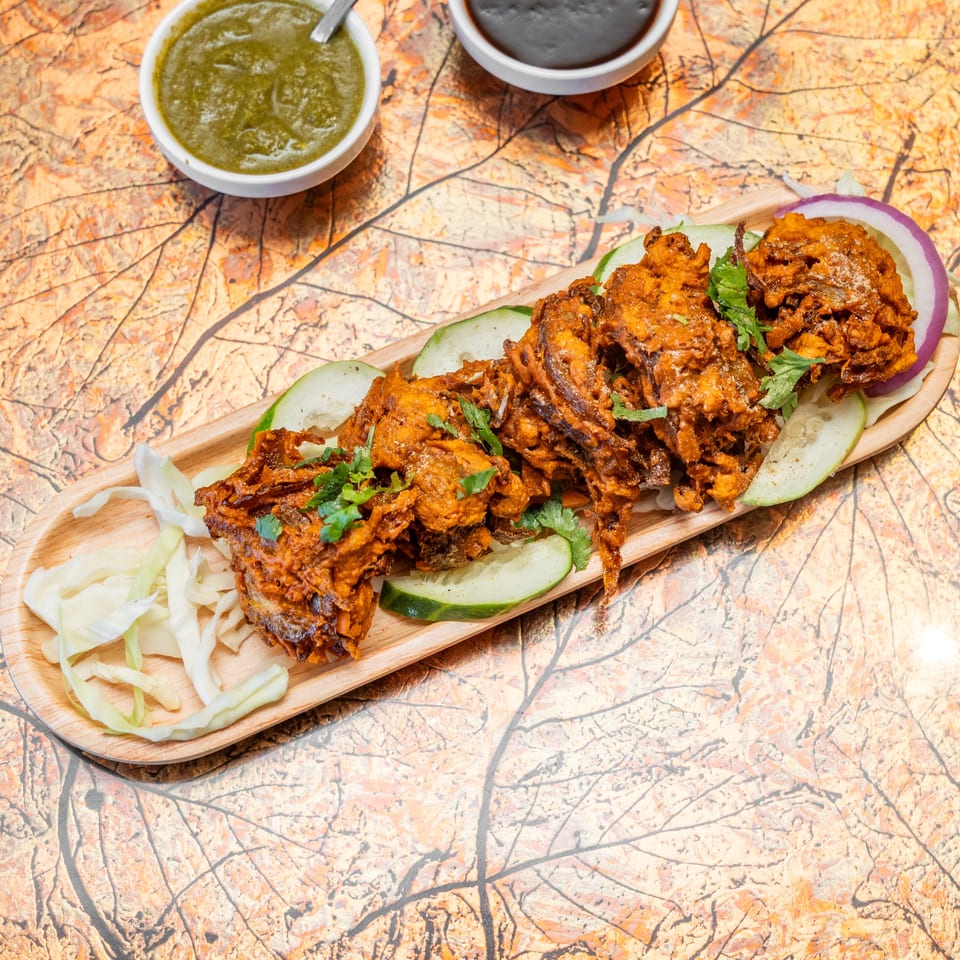 A wooden platter holds crispy spiced fritters garnished with cilantro, cucumber, cabbage, and onion, alongside green and dark dipping sauces.