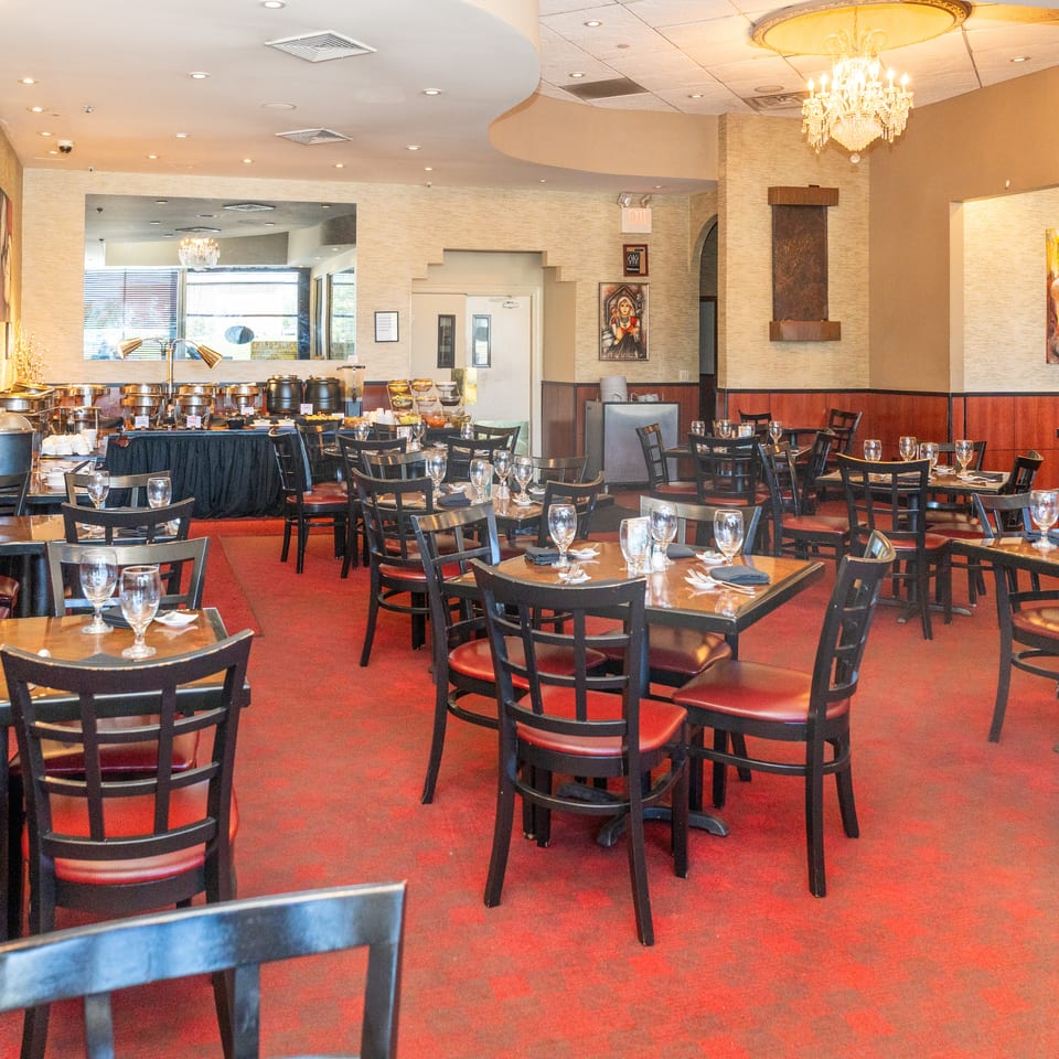 Elegant restaurant dining room with set tables, red-upholstered chairs, buffet station, chandeliers, and warm patterned walls.