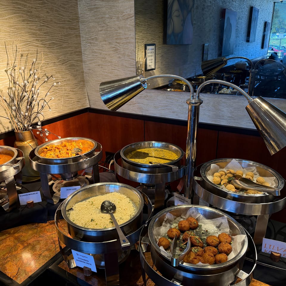 Indian buffet featuring chafing dishes of curries, rice, falafel, and fried appetizers beneath warming lamps.