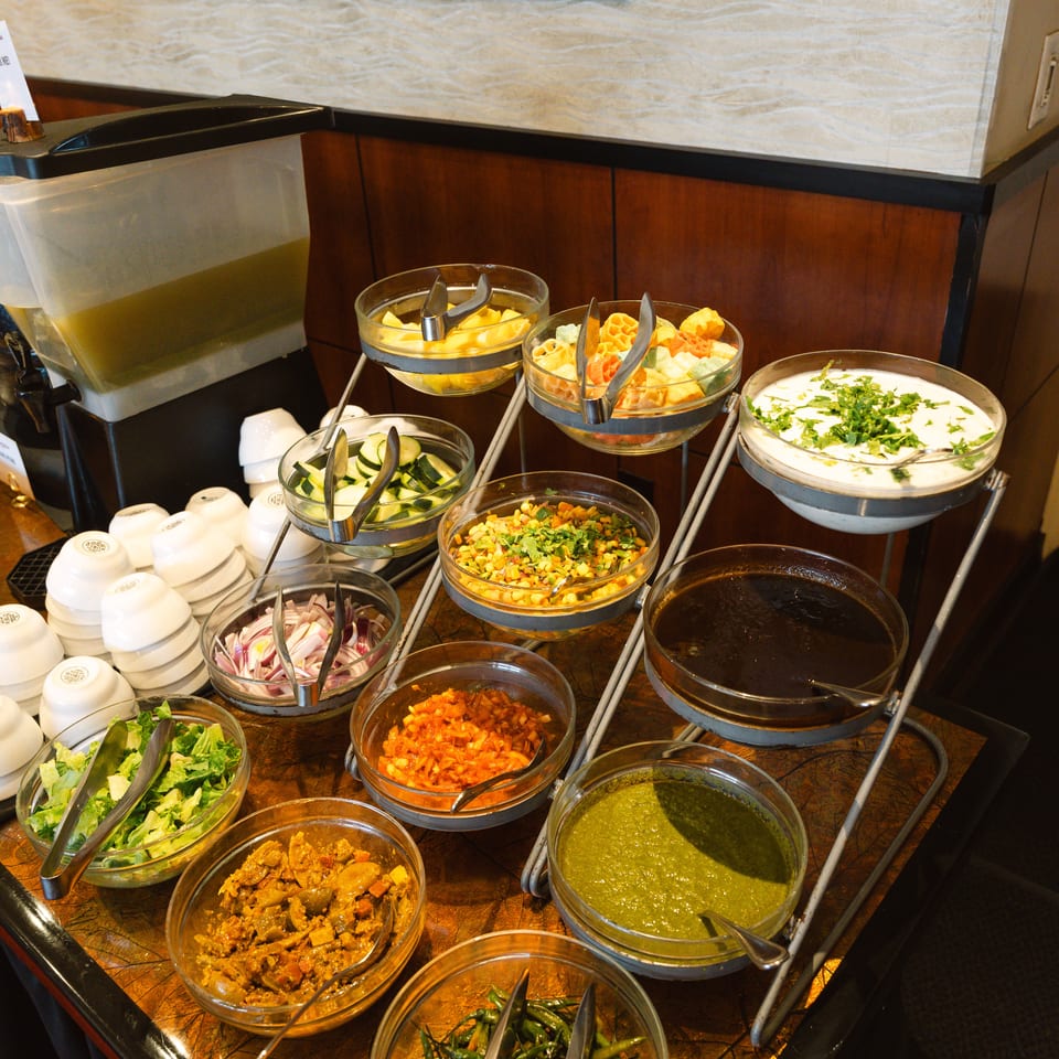 Buffet table displaying bowls of salads, vegetables, sauces, yogurt, and colorful toppings with serving utensils.