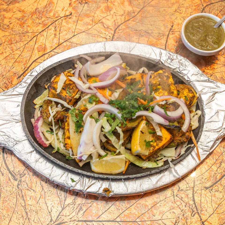 Steaming platter of grilled spiced meat and vegetables garnished with onions, cabbage, carrots, and cilantro, served with green chutney.