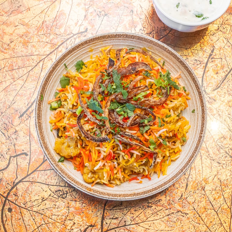 Colorful spiced rice topped with crispy fried vegetables and cilantro, served with creamy herb yogurt on a patterned table.