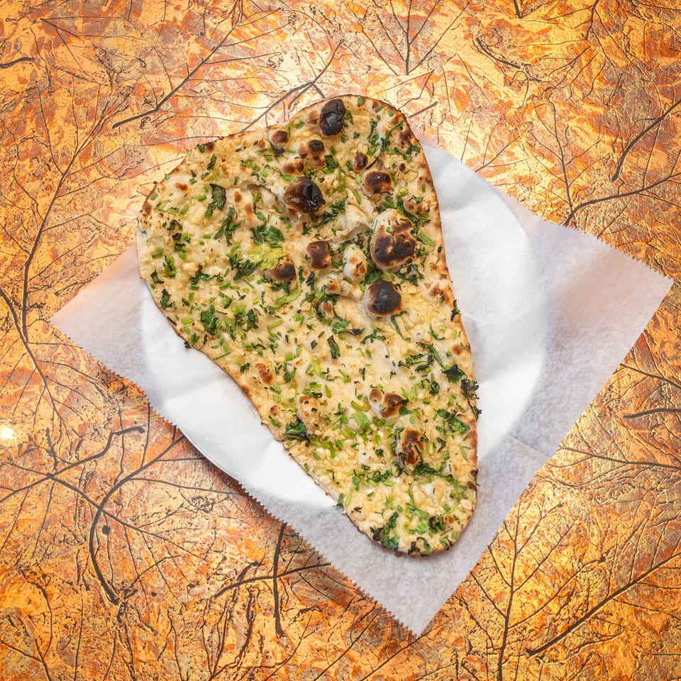 Garlic naan topped with chopped herbs and charred spots, served on parchment over an autumn leaf-patterned surface.