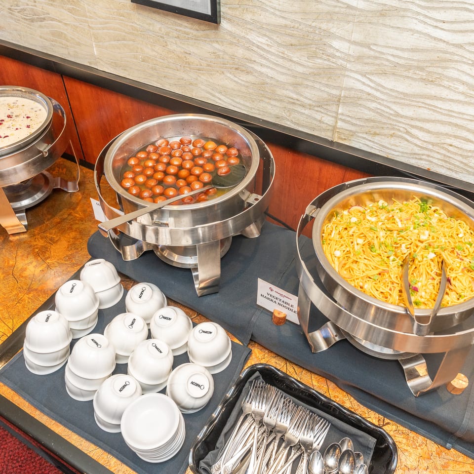Buffet station featuring gulab jamun, creamy dessert, vegetable hakka noodles, serving utensils, and stacked bowls.